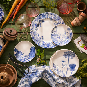 A collection of Caskata's Summer Blues Small Plates featuring floral and wildlife themes are displayed on a green wooden surface, surrounded by gardening tools, glassware, and vegetables.