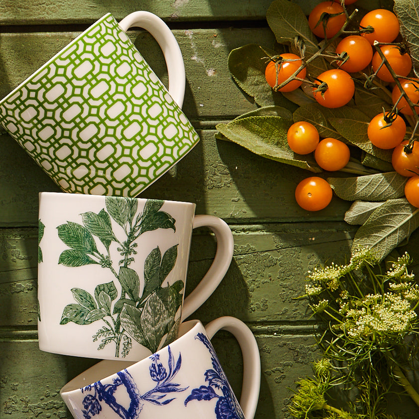 Three Caskata Arbor Green Mugs, featuring patterns of green geometric, botanical, and blue floral designs, all crafted from lead-free porcelain in Sri Lanka. They are beautifully arranged on a rustic green wooden surface with a sprig of yellow berries and lacy white flowers. These mugs are dishwasher safe for easy cleaning.