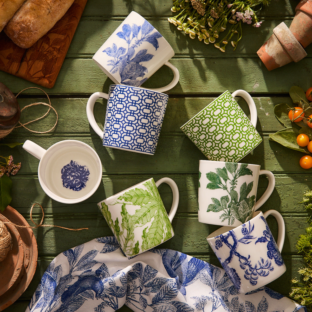 Six Arbor Mugs from Caskata Artisanal Home, crafted from premium porcelain and featuring blue and green leaf and geometric patterns, are beautifully arranged on a green wooden table amidst bread, flowers, and gardening tools. These charming ceramic mugs are both dishwasher and microwave-safe, making them a delightful addition to your kitchenware collection.