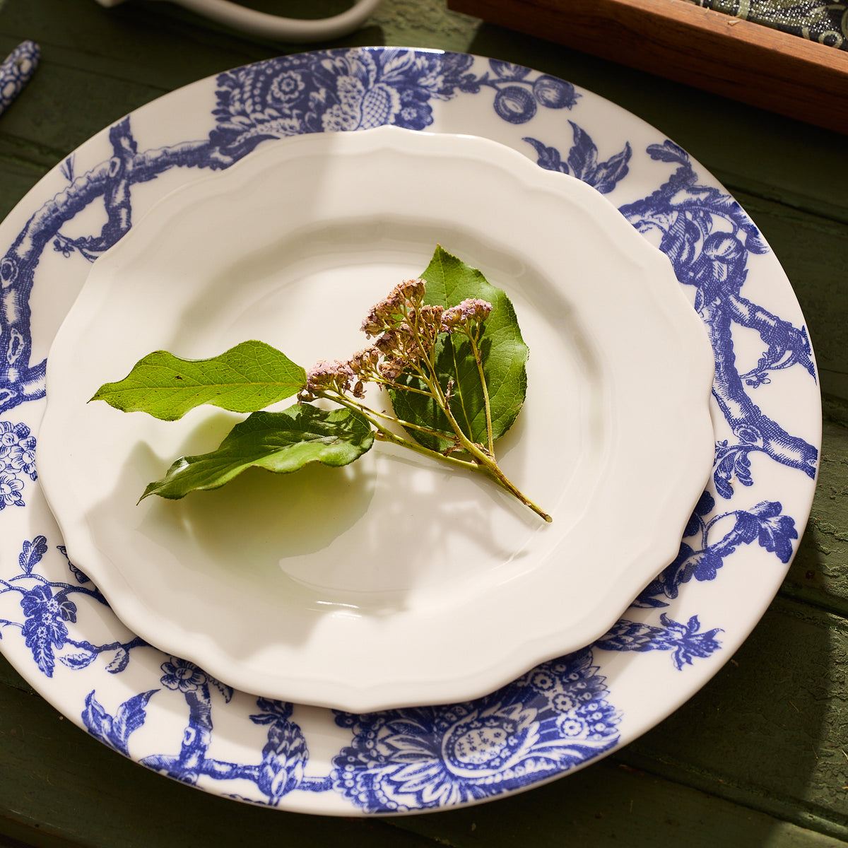 An Arcadia Rimmed Dinner Plate by Caskata Artisanal Home is placed on a table, with a white and blue decorative plate beneath it. On the top plate, there are a few green leaves and small, dried pink flowers. This display showcases premium porcelain dinnerware that elevates your dining experience with graphic florals and timeless fashion.