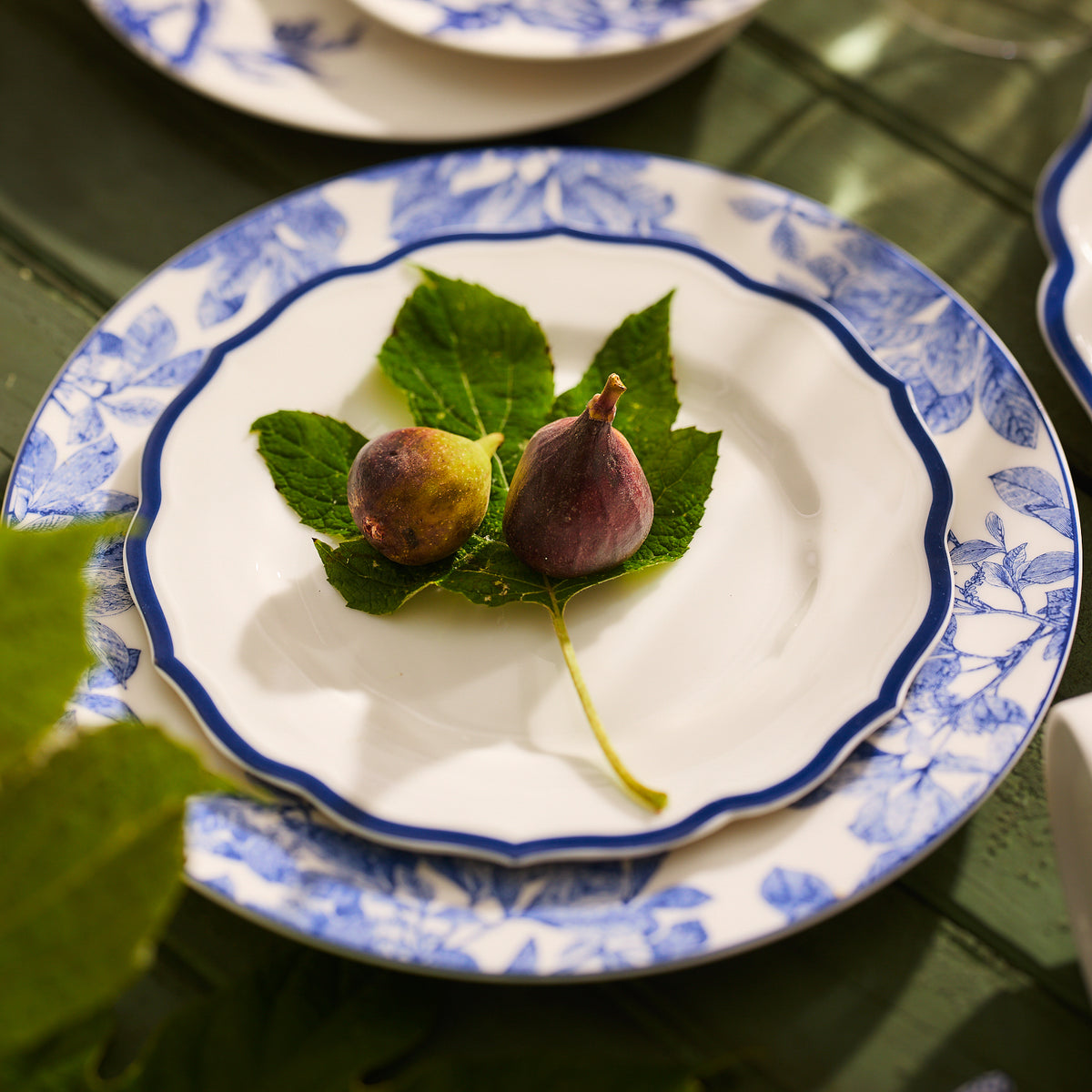 Two figs rest on a fig leaf, artfully centered on a decorative Stella Scalloped Blue Salad Plate by Caskata.