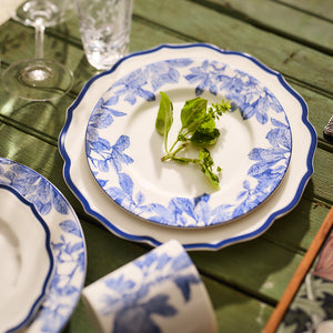 The Stella Scalloped Blue Dinner Plate from Caskata, featuring a charming heirloom-quality floral pattern in blue and white, holds a basil sprig on a green wooden table. Nearby sits a cup and glass, creating an inviting vintage ambiance.