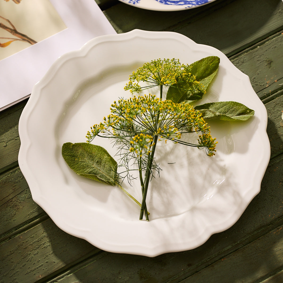 The High Tide Essentials Bundle by Caskata includes a white plate with green leaves and yellow flowers on a green wooden surface, ideal for pairing with school of fish napkins.