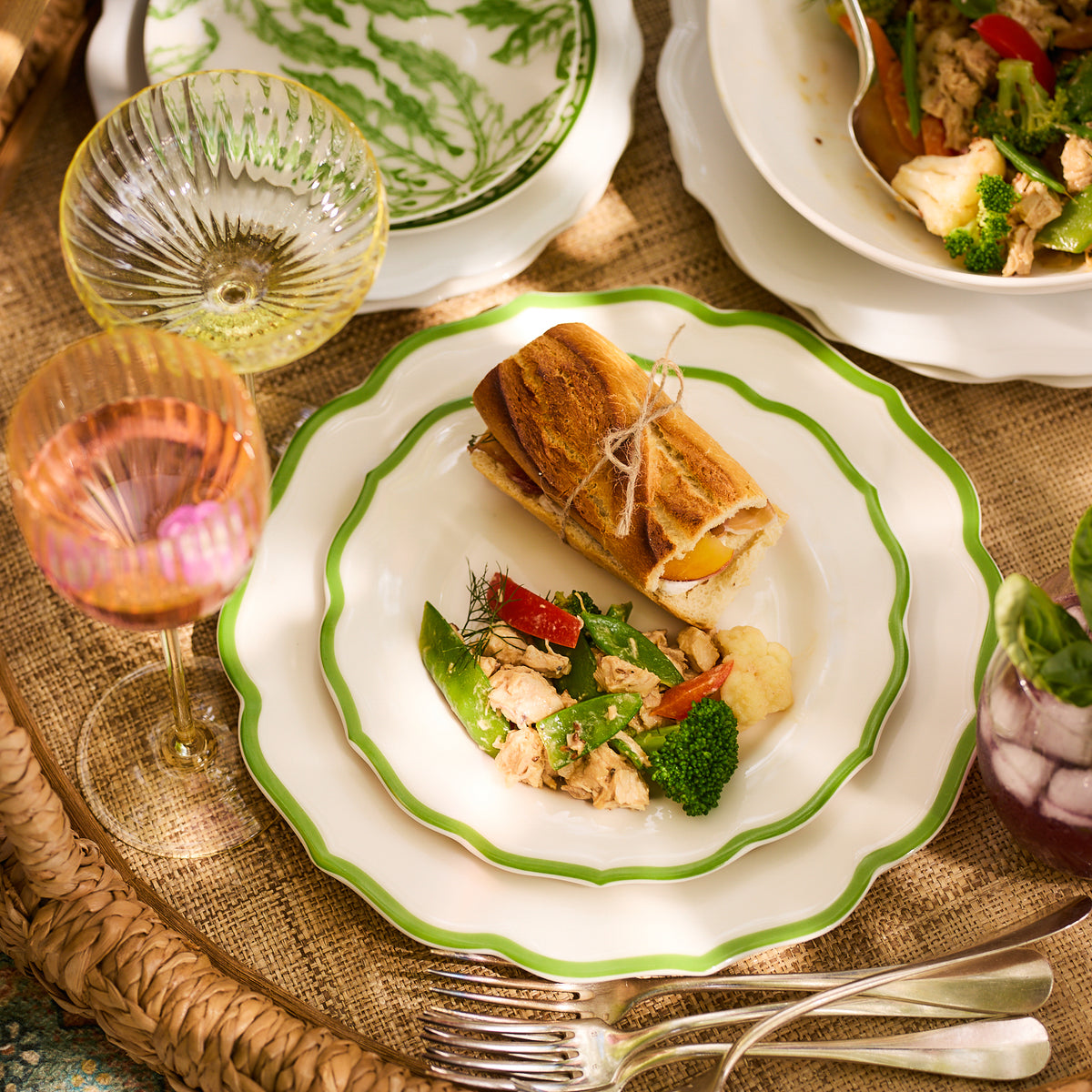 A table set with a plate of salad on the heirloom-quality Caskata Stella Scalloped Verde Dinner Plate sits alongside a bread roll and beverages in colorful glasses on a woven mat.