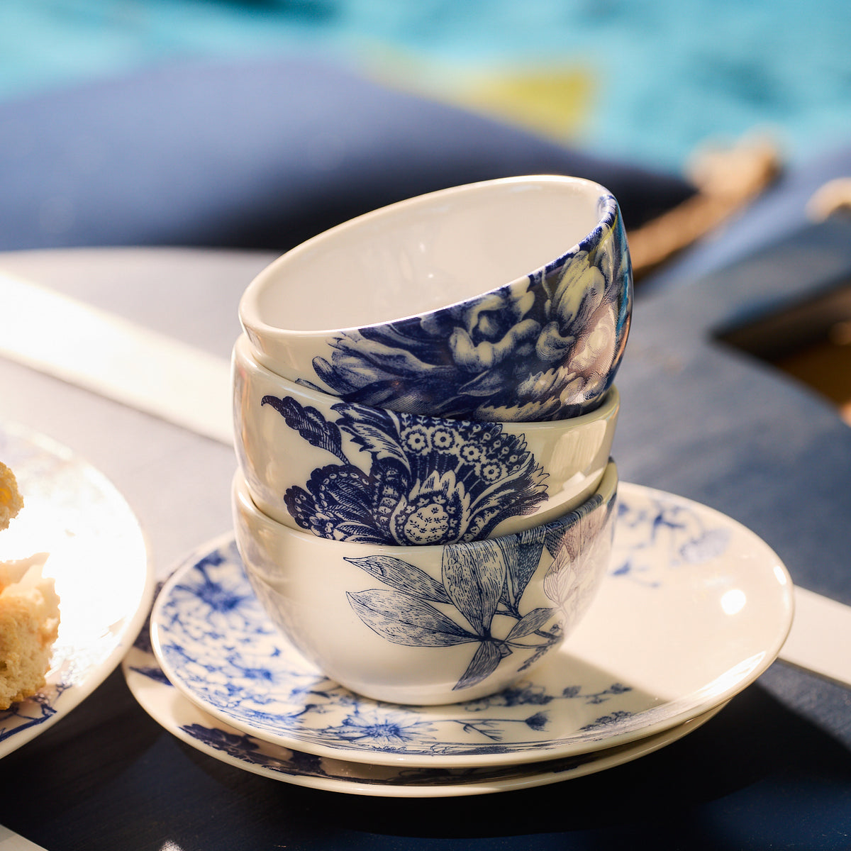 Three Arbor Snack Bowls from Caskata Artisanal Home, featuring blue floral designs nestled together inside a matching saucer, are placed on a dark blue table. Sunlight highlights the intricate patterns of the premium porcelain bowls, resembling leafy branches in delicate detail.