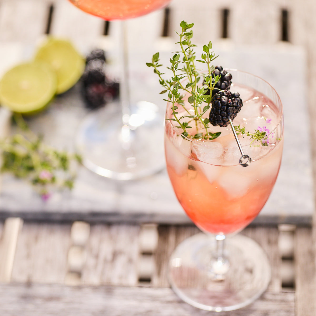 A cocktail in a Quinn Clear Everyday Glass by Caskata Artisanal Home, garnished with a blackberry, thyme, and ice cubes, sits on a wooden table. Slices of lime and another drink are blurred in the background. The lead-free crystal glass adds an elegant touch to this scene from the Czech Republic.