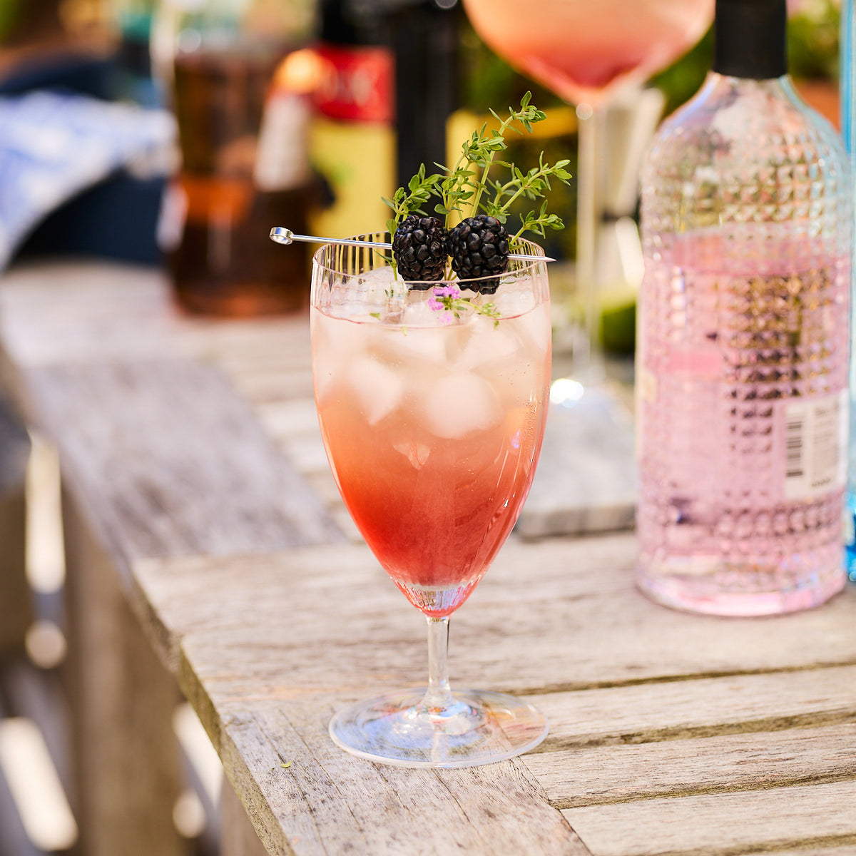 A Quinn Clear Everyday Glass by Caskata Artisanal Home, mouth-blown and crafted from lead-free crystal, is filled with a pinkish drink, ice cubes, and garnished with blackberries and a sprig of greenery. The exquisite Czech Republic glassworks piece rests on a wooden table amidst other bottles and glasses in the background.