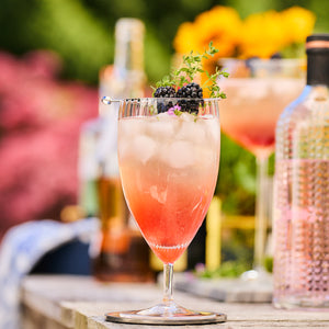 A glass of iced pink cocktail, garnished with blackberries and a sprig of thyme, sits on a table outdoors. The exquisite Quinn Clear Everyday Glasses by Caskata Artisanal Home add elegance to the scene, with bottles and a second mouth-blown glass blurred in the background.