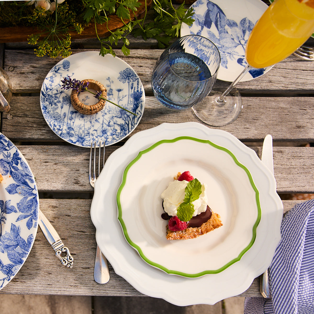 A slice of cake topped with cream and raspberries graces a dessert plate from Caskata&#39;s Stella Scalloped Verde collection on an outdoor wooden table. The floral plates, cutlery, glass, and orange drink complement the neutral palette, adding an heirloom feel to this charming setting.