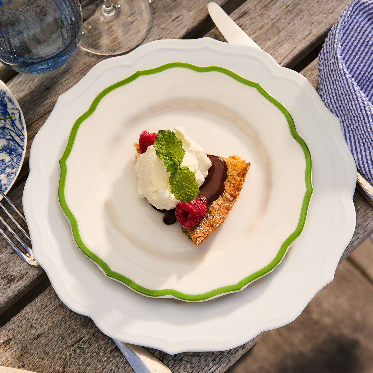 A dessert presented on a Stella Scalloped Verde Salad Plate from Caskata, showcasing a slice of cake adorned with whipped cream, chocolate sauce, raspberries, and a mint leaf. The white plate with its green rim offers an heirloom feel as it sits gracefully on the wooden table.