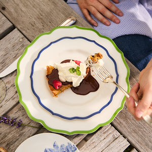 A person enjoys a dessert topped with chocolate sauce and whipped cream on a Stella Scalloped Blue Salad Plate by Caskata, using a fork. The wooden table adds warmth, complemented by a nearby lavender sprig that enhances the scene's charm.