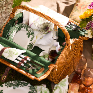 A wicker picnic basket featuring Arbor Green Rimmed Salad Plates by Caskata, along with premium cutlery, napkins, a bottle of wine, and two glasses set on a blanket next to a small floral bouquet.