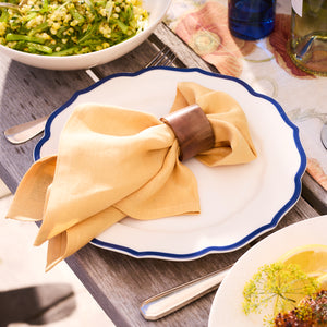 A yellow cloth napkin with a wooden ring rests on the Stella Scalloped Blue Dinner Plate by Caskata, exuding a vintage feel on the wooden table next to a bowl of salad.