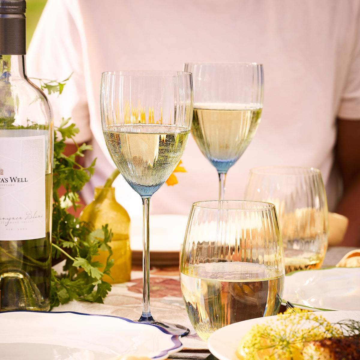 A table setting with a person in a pink shirt, three Quinn Ocean White Wine Glasses from Caskata Artisanal Home filled with white wine, a bottle of white wine, a carafe, and plates with food. A sprig of fresh herbs is placed next to the wine bottle. The elegant lead-free Quinn Ocean White Wine Glasses add a touch of sophistication.