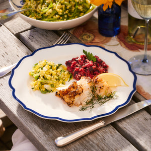 The beautifully presented dish of grilled fish with a lemon wedge, accompanied by corn salad and beet salad, is elegantly garnished with dill and served on a Stella Scalloped Blue Dinner Plate from Caskata. The wooden table enhances the vintage aura of this meal.