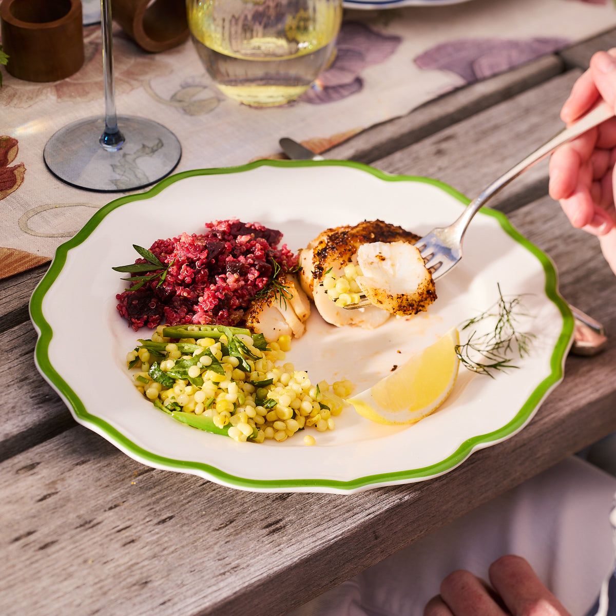 Using the Stella Scalloped Verde Dinner Plate by Caskata, a dish featuring seared scallops, red quinoa salad, corn, greens, and a lemon wedge is elegantly presented. A hand with a fork hovers above the meal.