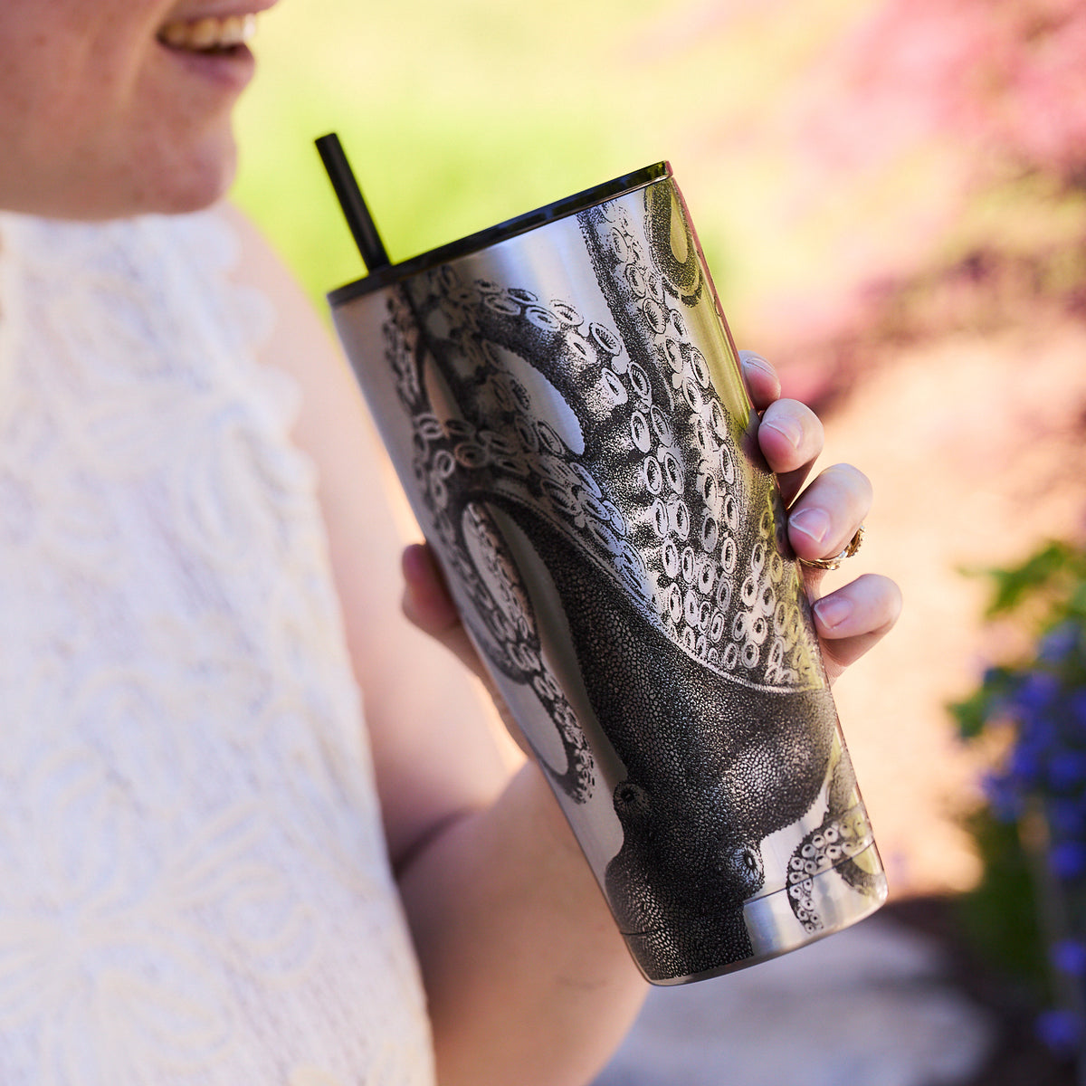 A person enjoying their drink from a Caskata Lucy Chrome Stainless Steel Insulated Tumbler, the stainless steel gleaming with an intricate cephalopod design, as they sip through a black straw.