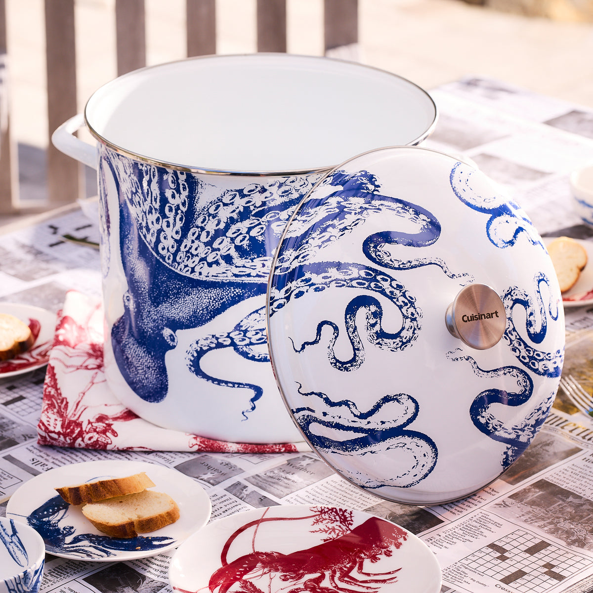 The Caskata X Cuisinart Lucy 16 Qt. Enamel on Steel Stockpot, featuring a blue octopus design and accompanied by a matching lid, rests on a table covered with newspapers. Nearby are sandwiches and a lobster napkin, perfect for clambakes and more.