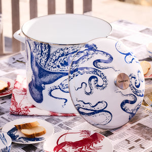 The Caskata X Cuisinart Lucy 16 Qt. Enamel on Steel Stockpot, featuring a blue octopus design and accompanied by a matching lid, rests on a table covered with newspapers. Nearby are sandwiches and a lobster napkin, perfect for clambakes and more.