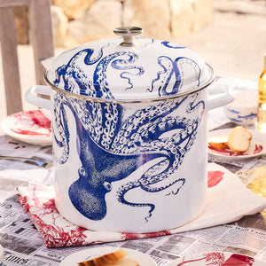 A large Caskata X Cuisinart Lucy 16 Qt. Enamel on Steel Stockpot, adorned with a blue octopus design, sits on a table covered with newspaper and assorted food items, perfect for clambakes.