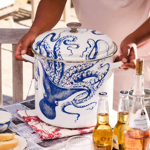 Person holding a large Caskata X Cuisinart Lucy 16 Qt. Enamel on Steel Stockpot featuring a blue octopus design at a table adorned with alcoholic beverages and food, perfect for clambakes.