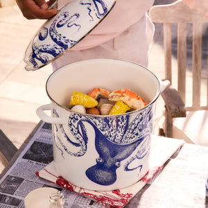 A person lifts the lid off a Caskata X Cuisinart Lucy 16 Qt. Enamel on Steel Stockpot, featuring a blue octopus design from Cuisinart. The pot, ideal for clambakes, contains corn, clams, and crabs and rests on newspapers covering a table.