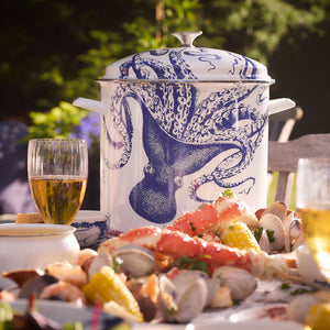 A seafood boil feast spread on a table with clams, corn, and crab legs. A large white Caskata X Cuisinart Lucy 16 Qt. Enamel on Steel Stockpot with a blue octopus design and two glasses of beer are also visible, evoking the charm of traditional clambakes.