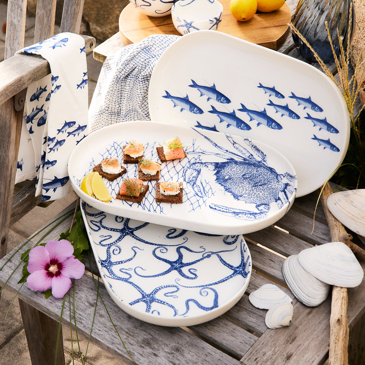 The &quot;School of Fish Large Coupe Oval Platter&quot; by Caskata, crafted from high-fired porcelain and featuring intricate designs of blue fish, octopus, and fishing nets, is elegantly arranged outdoors. This platter with raised rims skillfully holds small appetizers alongside a lemon slice. Scattered leaves and flowers add to the enchanting ambiance of this charming fish dinnerware display.