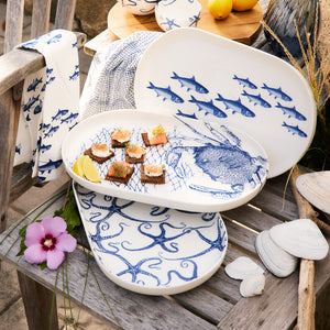 Caskata's Starfish Large Coupe Oval Platter with blue marine designs of fish and octopuses rests elegantly on a wooden chair. Some platters display appetizers alongside a lemon wedge and a purple flower. Nearby, seashells, starfish, and a towel create an ideal setting for ocean-themed entertaining.