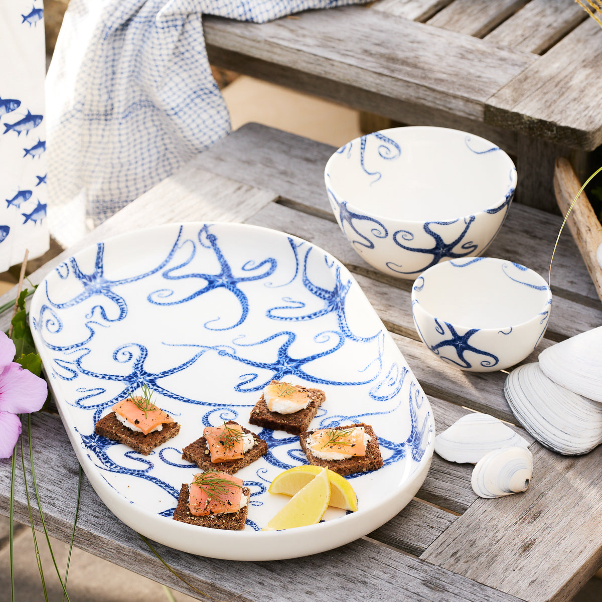 A Starfish Snack Bowl from Caskata Artisanal Home, adorned with intricate blue octopus designs, sits on a wooden table filled with hors d&#39;oeuvres and lemon wedges. Nearby, seashell and starfish decorations enhance the scene alongside a folded cloth with blue patterns.