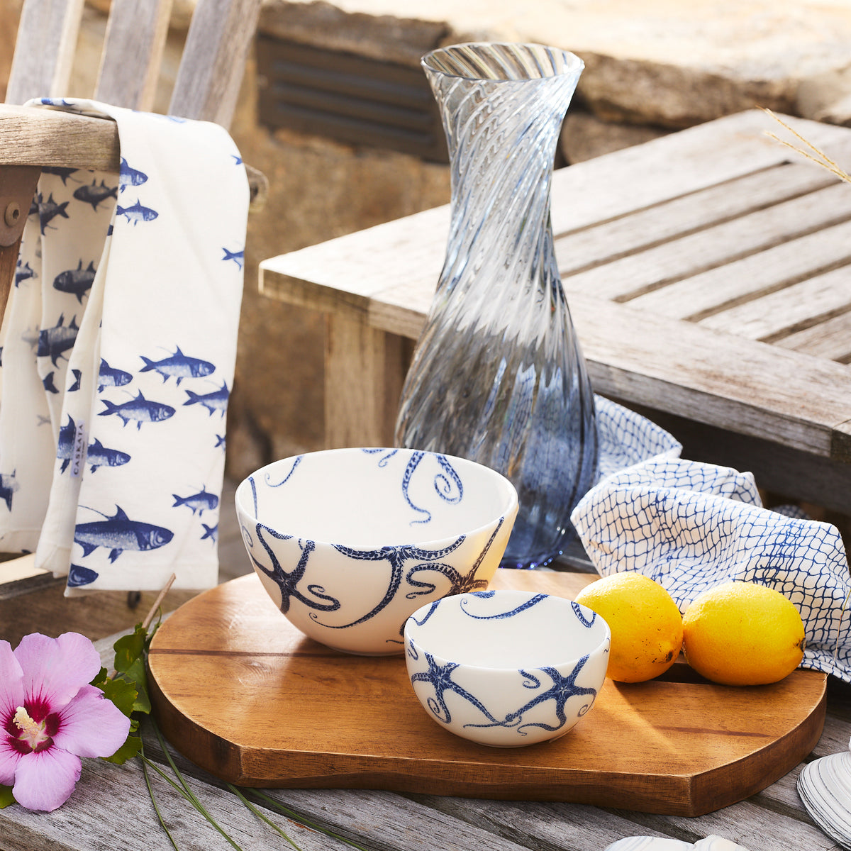 The Starfish Snack Bowls from Caskata Artisanal Home, featuring elegant blue marine starfish designs, are placed on a wooden tray alongside a glass vase, lemons, a floral cloth, and a pink flower on the wooden table.