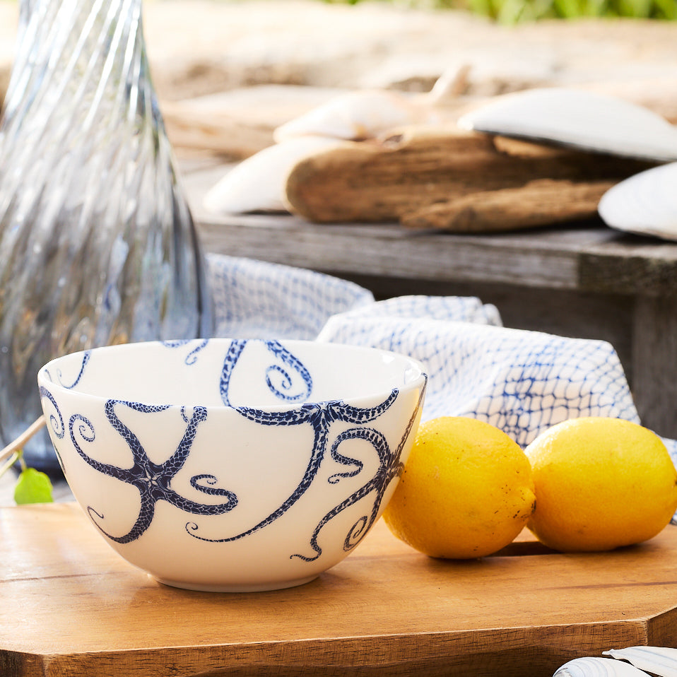 A Caskata Starfish Cereal Bowl, crafted from white porcelain and adorned with blue octopus designs, sits on a wooden surface next to two lemons.