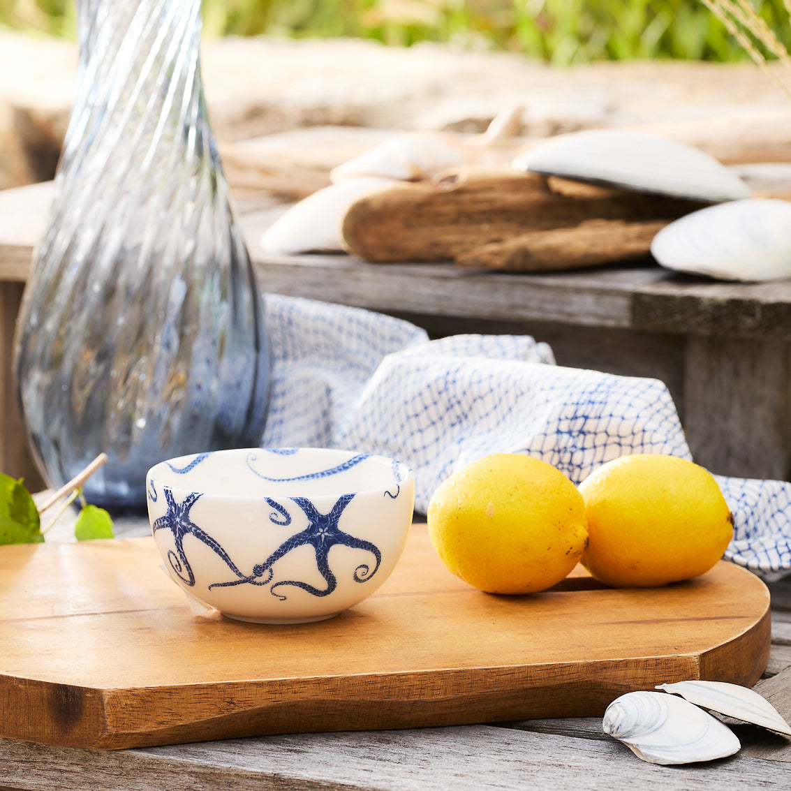 A Caskata Artisanal Home Starfish Snack Bowl containing two lemons rests on a wooden board. In the background, there&#39;s a glass vase alongside a cloth and seashells on a rustic wooden surface.