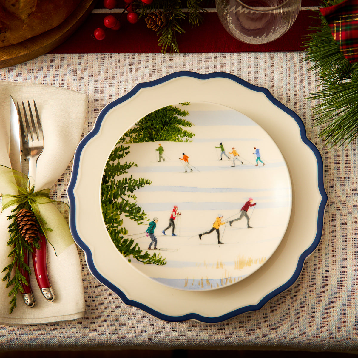 A festive table setting showcasing Caskata&#39;s heirloom-quality Stella Scalloped Blue Dinner Plate, complemented by a skiing-themed decorative plate and red-handled cutlery wrapped in a napkin adorned with a pinecone embellishment. The arrangement exudes a vintage feel perfect for the season.