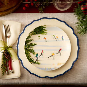 A festive table setting showcasing Caskata's heirloom-quality Stella Scalloped Blue Dinner Plate, complemented by a skiing-themed decorative plate and red-handled cutlery wrapped in a napkin adorned with a pinecone embellishment. The arrangement exudes a vintage feel perfect for the season.