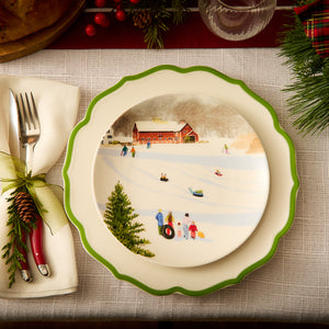 The Stella Scalloped Verde Dinner Plate from Caskata showcases a winter scene of people sledding, set on a green-rimmed plate with elegant scalloped details, complemented by cutlery wrapped in a napkin and accented with pine cones and greenery.