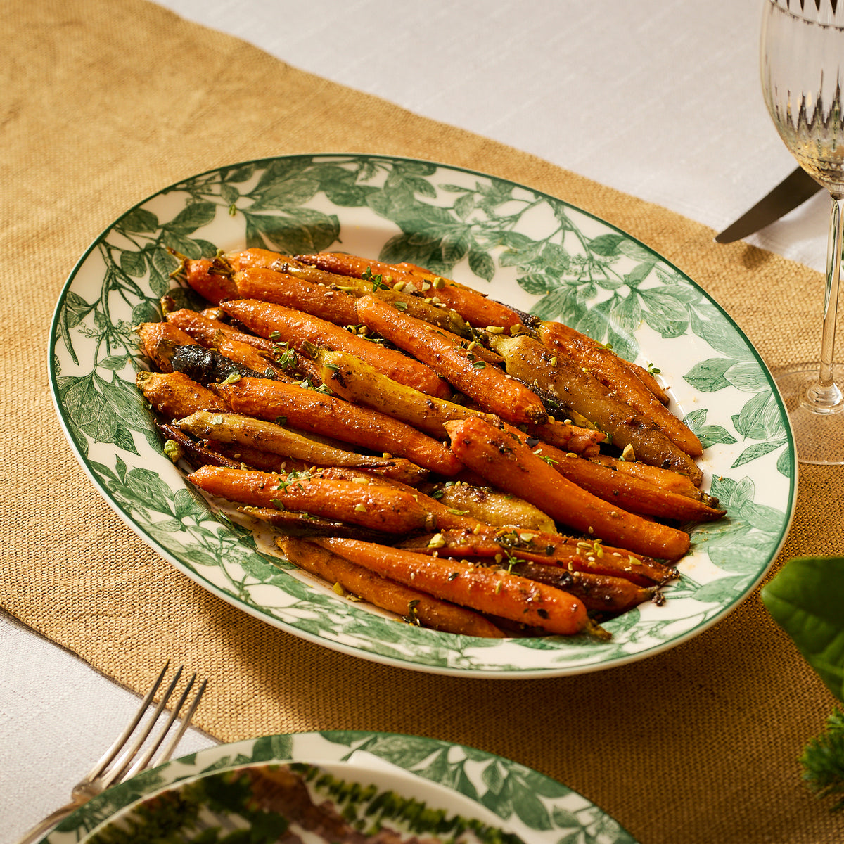 A plate of glazed carrots garnished with herbs is elegantly presented on the Arbor Green Oval Rimmed Platter from Caskata Artisanal Home, resting gracefully on a burlap runner with silverware nearby.