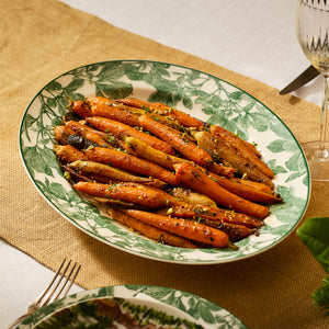 A plate of glazed carrots garnished with herbs is elegantly presented on the Arbor Green Oval Rimmed Platter from Caskata Artisanal Home, resting gracefully on a burlap runner with silverware nearby.