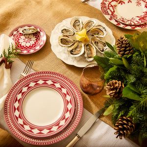 For a holiday table setting, feature the elegant Fez Crimson Rimmed Salad Plates from Caskata Artisanal Home in a timeless shape, complemented by a napkin adorned with a pine cone and greenery. Add a plate of oysters with lemon, utensils, and a glass on a burlap table runner.