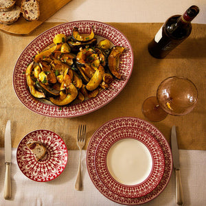 A set table with Casablanca Crimson Rimmed Dinner Plate by Caskata Artisanal Home, featuring ornate scrollwork, a serving platter of roasted vegetables, a bottle of red wine, an empty wine glass, cutlery, and sliced bread on a wooden board.