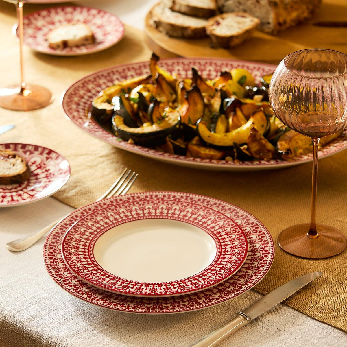 Table setting with red-patterned plates, cutlery, and a glass. A large porcelain dinner plate with a roasted vegetable dish is at the center. Bread slices in the background. White tablecloth and beige table runner add elegance to the scene, complemented by Caskata Artisanal Home&#39;s Casablanca Crimson Rimmed Dinner Plate&#39;s ornate scrollwork.