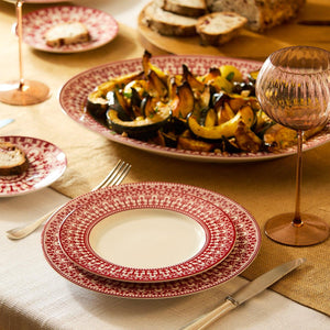 Table setting with red-patterned plates, cutlery, and a glass. A large porcelain dinner plate with a roasted vegetable dish is at the center. Bread slices in the background. White tablecloth and beige table runner add elegance to the scene, complemented by Caskata Artisanal Home's Casablanca Crimson Rimmed Dinner Plate's ornate scrollwork.