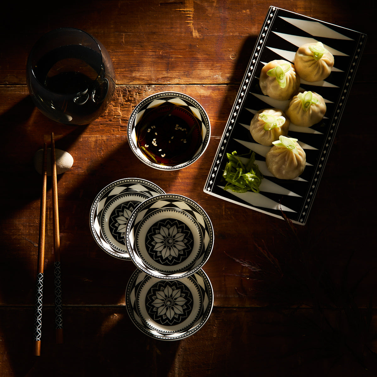 A table showcasing Caskata&#39;s Fez Dipping Dishes set with intricate patterns, alongside patterned plates, a glass, chopsticks, and a rectangular plate with dumplings and greens embodies Morocco&#39;s vibrant essence.