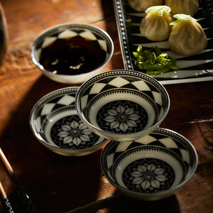 A set of four Caskata Fez Dipping Dishes, featuring striking black and white geometric patterns, sits beside a plate of dumplings and chopped greens on a wooden table, evoking artistic flair reminiscent of Morocco.