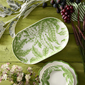 A Freya Large Coupe Oval Platter by Caskata, featuring green floral patterns, sits on a green wooden table alongside grapes, carrots, and flowers—an essential for entertaining.