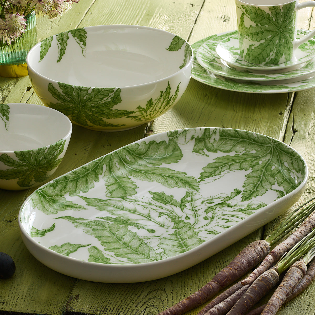 The Freya Vegetable Serving Bowl from Caskata Artisanal Home is a piece of white ceramic tableware adorned with delicate green florals, beautifully showcased on a wooden surface.