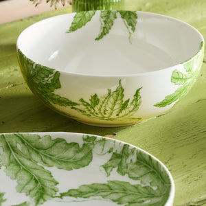 A Freya Vegetable Serving Bowl from Caskata Artisanal Home, crafted in white porcelain and adorned with green floral patterns, rests gracefully on a textured green surface.