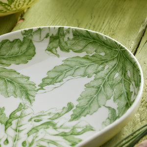 The Freya Large Coupe Oval Platter by Caskata, featuring a green leafy pattern, rests elegantly on a textured green wooden surface, making it an entertaining must-have for any gathering.