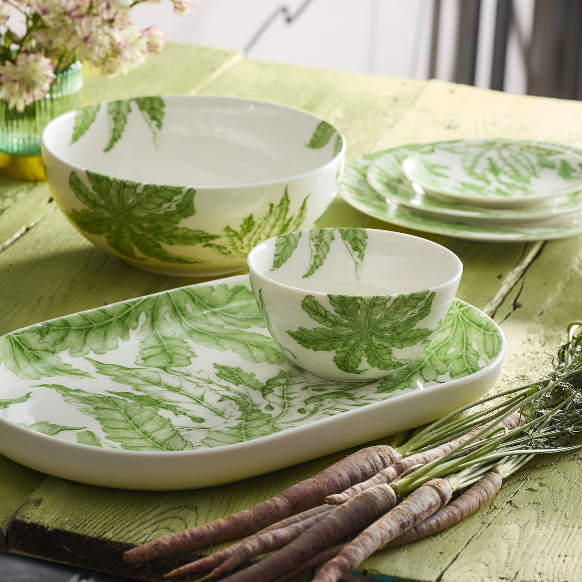 The ceramic dinnerware set, featuring the dishwasher and microwave safe Freya Cereal Bowls from Caskata adorned with green floral porcelain patterns, is elegantly displayed on a green wooden table. Alongside lies a bunch of fresh carrots and a small flower arrangement.