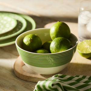 A Newport Stripe Verde Cereal Bowl from Caskata sits on a wooden surface, accompanied by a matching napkin. A sliced lime and a glass of ice water glisten in the background, while green plates crafted from high-fired porcelain are neatly stacked to the side.